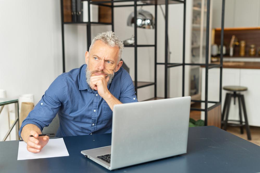 Absorbed pensive senior businessman in smart casual shirt sits at the desk and using laptop, lost in thoughts mature grey hair man looking for new ideas, waiting for inspiration
